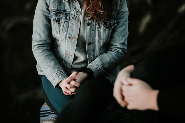 Grieving a cat can sometimes require professional help like the therapy session depicted in this photo. A person with long hair in a jean jacket is sitting with her hands crossed while another person, slightly blurred, wearing black has their crossed hands visible.