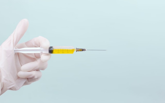 A light blue background with a white gloved hand on the left side. The hand is holding a needle and syringe with a yellow liquid in it.