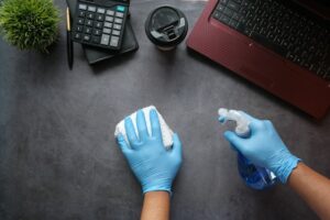 A person's hands with blue disposable gloves, a rag, and a squirt bottle clean a desk with a red laptop, disposable coffee cup, calculator, and plant on it.