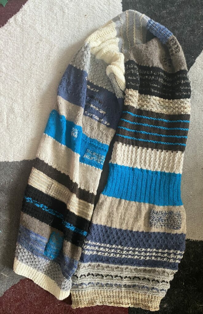 A blue, gray, cream, brown, and black colored knit scarf sits on a rug. The colors are in stripes with differing textures and are the same color as Zoloft, Joey's beloved cat.