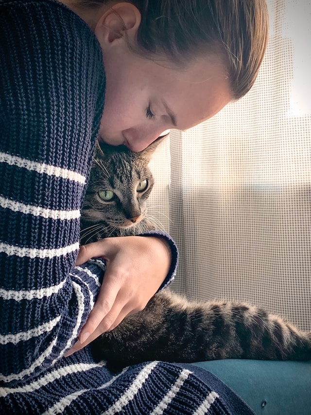 A woman in a navy blue sweater with white stripes holds a brown tabby cat with green eyes in her lap.