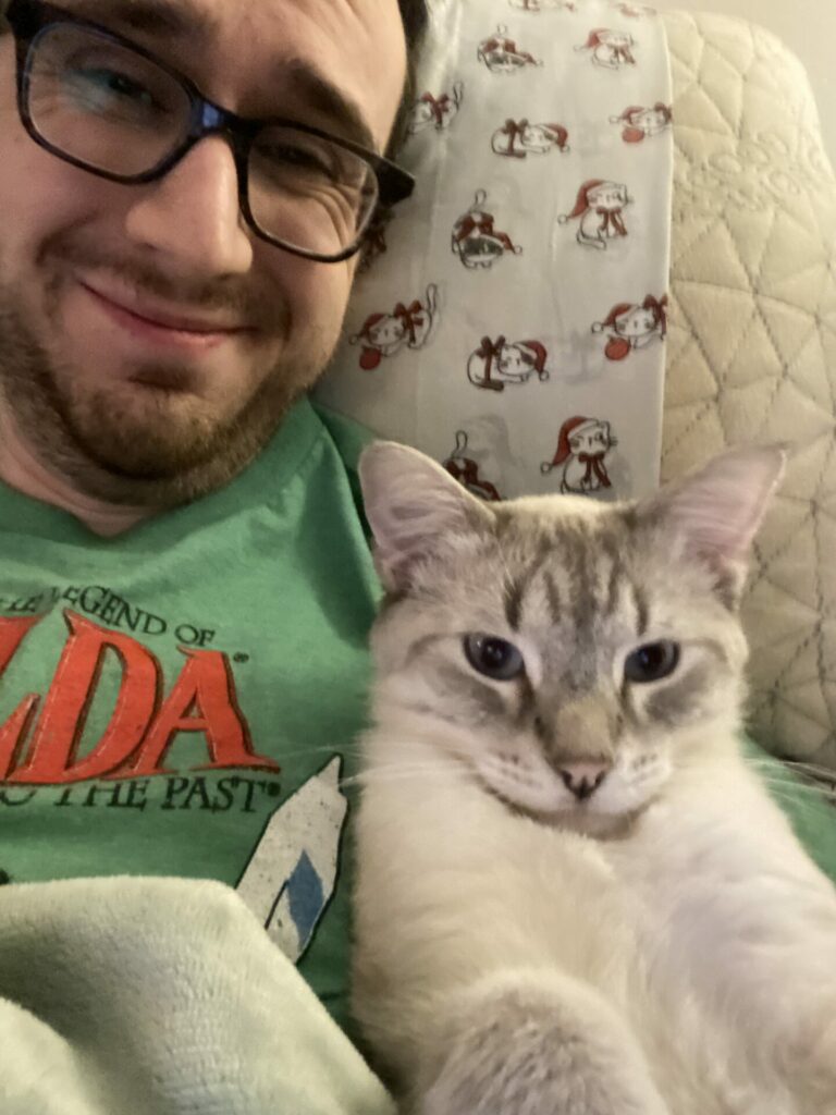 Joey, a white male with glasses, dark hair, and a short beard, lays in bed while wearing a green shirt with Poutine, a lynx point Siamese cat.
