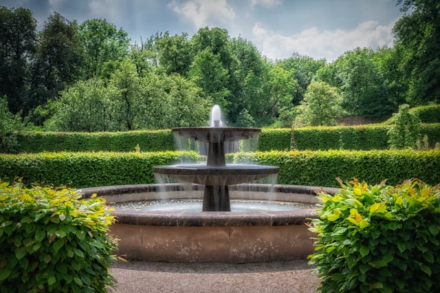 A stone water fountain with two basins and a small geyser in the middle is seen in the middle of a stone pool. There are green hedges and partially cloudy skies behind it. It appears to be in a park.