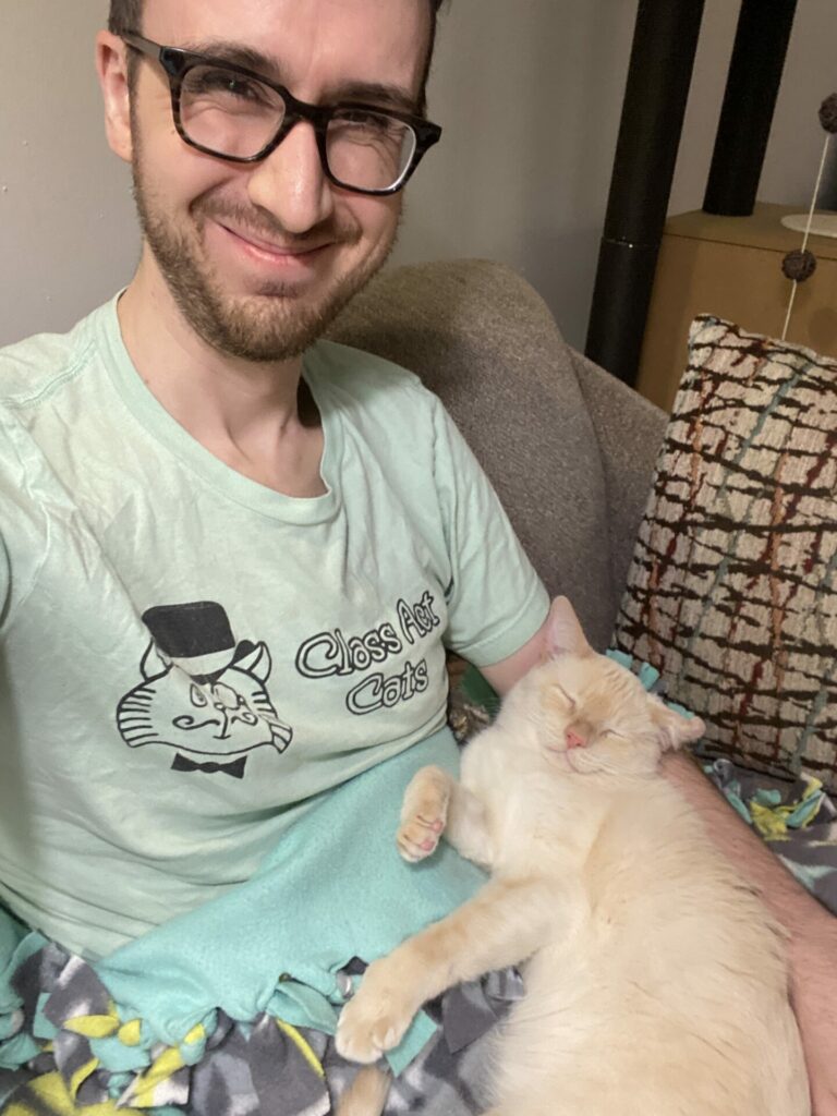 Joey is wearing a mint Class Act Cats shirt and is smiling sitting on a couch. Prozac, a flame point Siamese cat, is sleeping on his lap with his arms in a goofy position curled into Joey's arm.